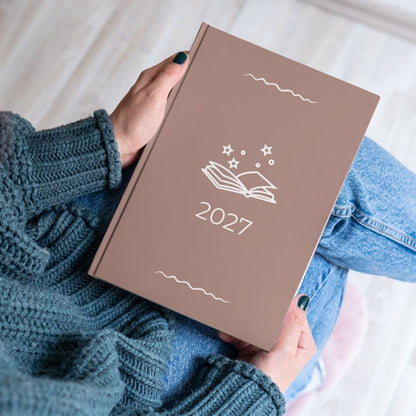 Earthy mushroom diary with a white open book and stars outline titled '2027' held by the hands of a women in a blue knitted jumper and jeans. 
