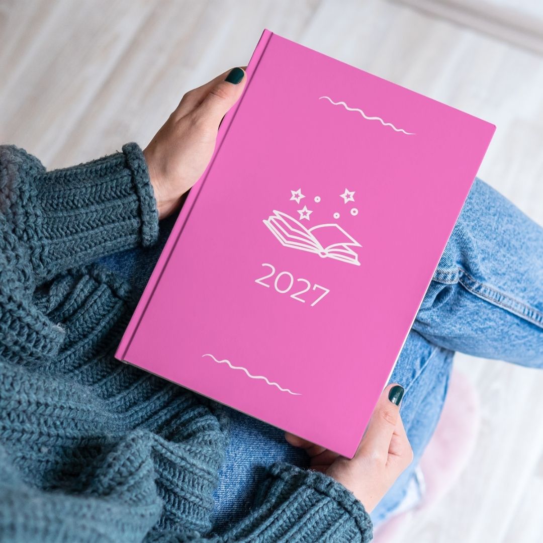 Flamingo pink diary with a white open book and stars outline titled '2027' held by the hands of a women in a blue knitted jumper and jeans. 
