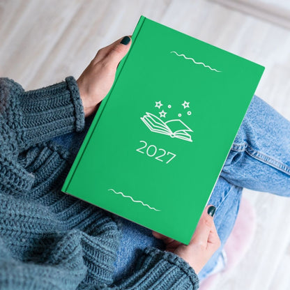Forest green diary with a white open book and stars outline titled '2027' held by the hands of a women in a blue knitted jumper and jeans. 