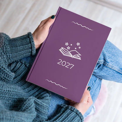 Plum purple diary with a white open book and stars outline titled '2027' held by the hands of a women in a blue knitted jumper and jeans. 