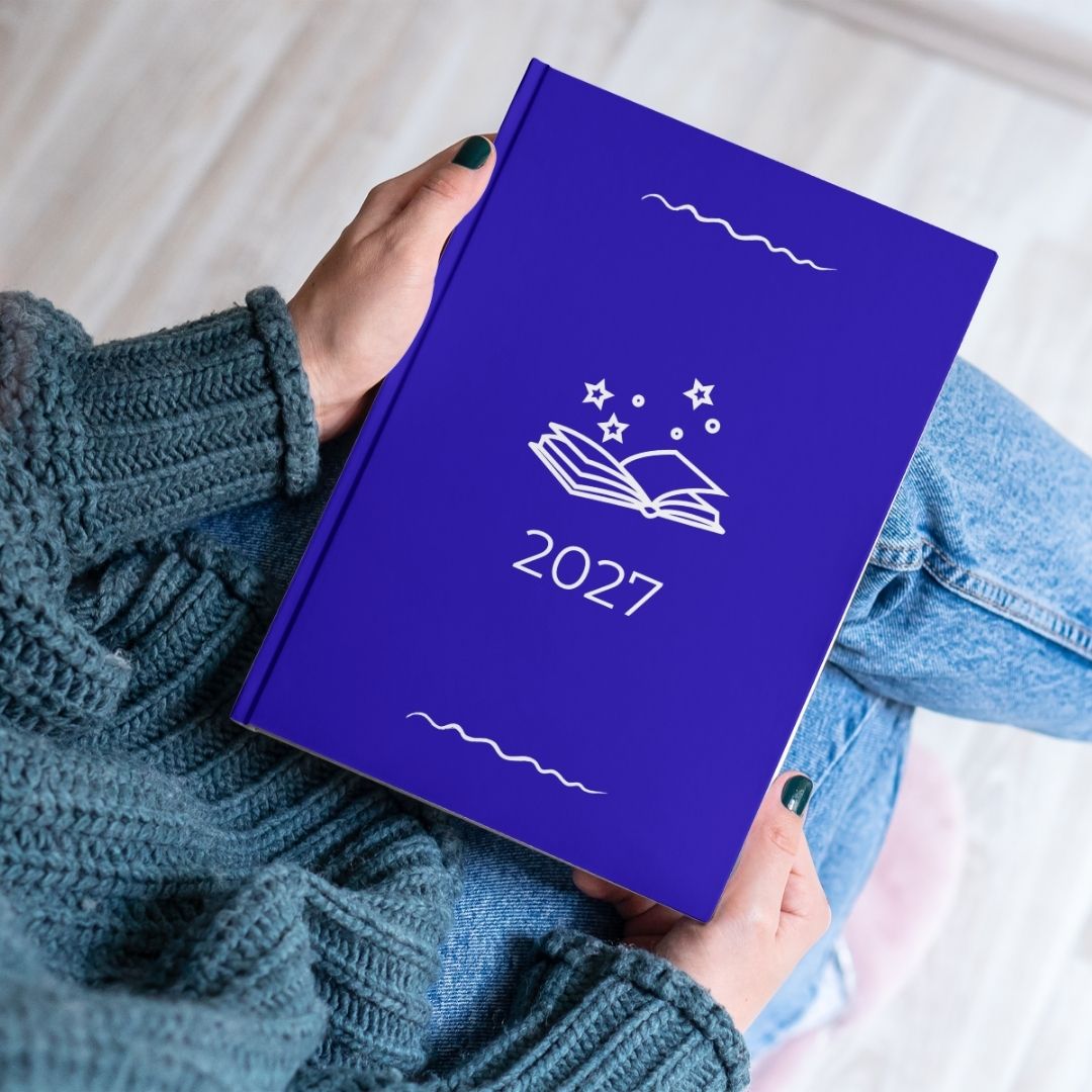 Royal blue diary with a white open book and stars outline titled '2027' held by the hands of a women in a blue knitted jumper and jeans. 