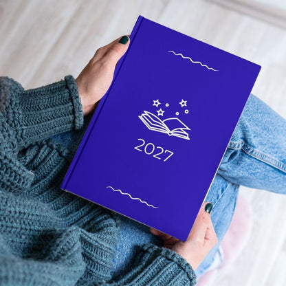 Royal blue diary with a white open book and stars outline titled '2027' held by the hands of a women in a blue knitted jumper and jeans. 