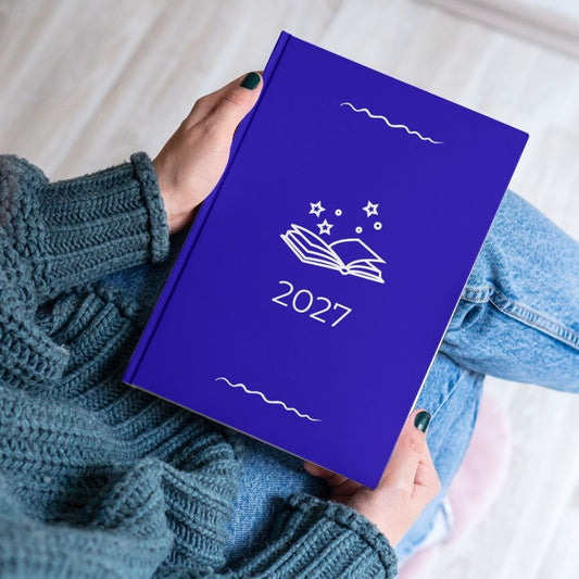 Royal blue diary with a white open book and stars outline titled '2027' held by the hands of a women in a blue knitted jumper and jeans. 