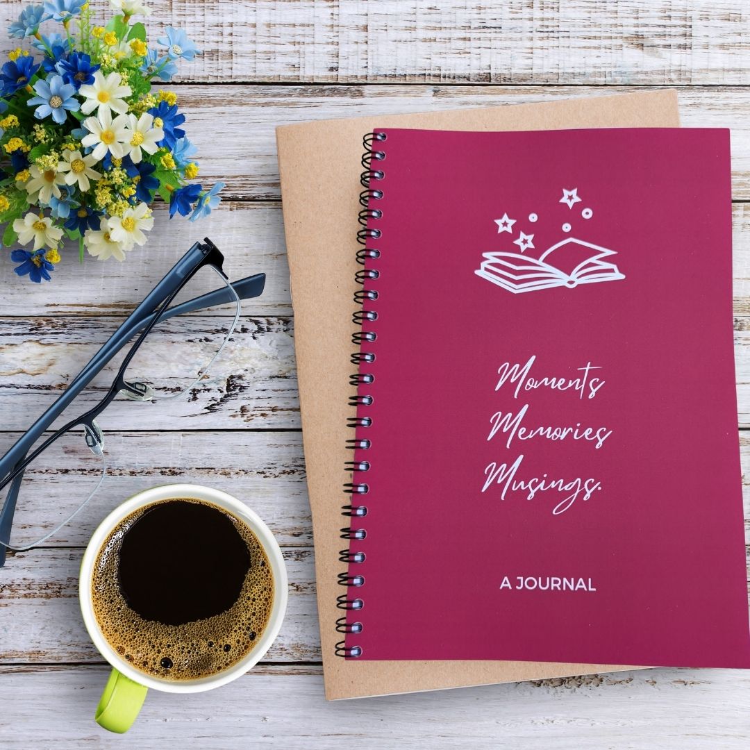 A journal with a solid magenta background and white line art of an open book with stars above the title 'Moments Memories Musings' and 'A JOURNAL' text at the bottom. The journal sits on a bleached wooden table besides a mug of coffee, pair of glasses, and blue white green flower arrangement.