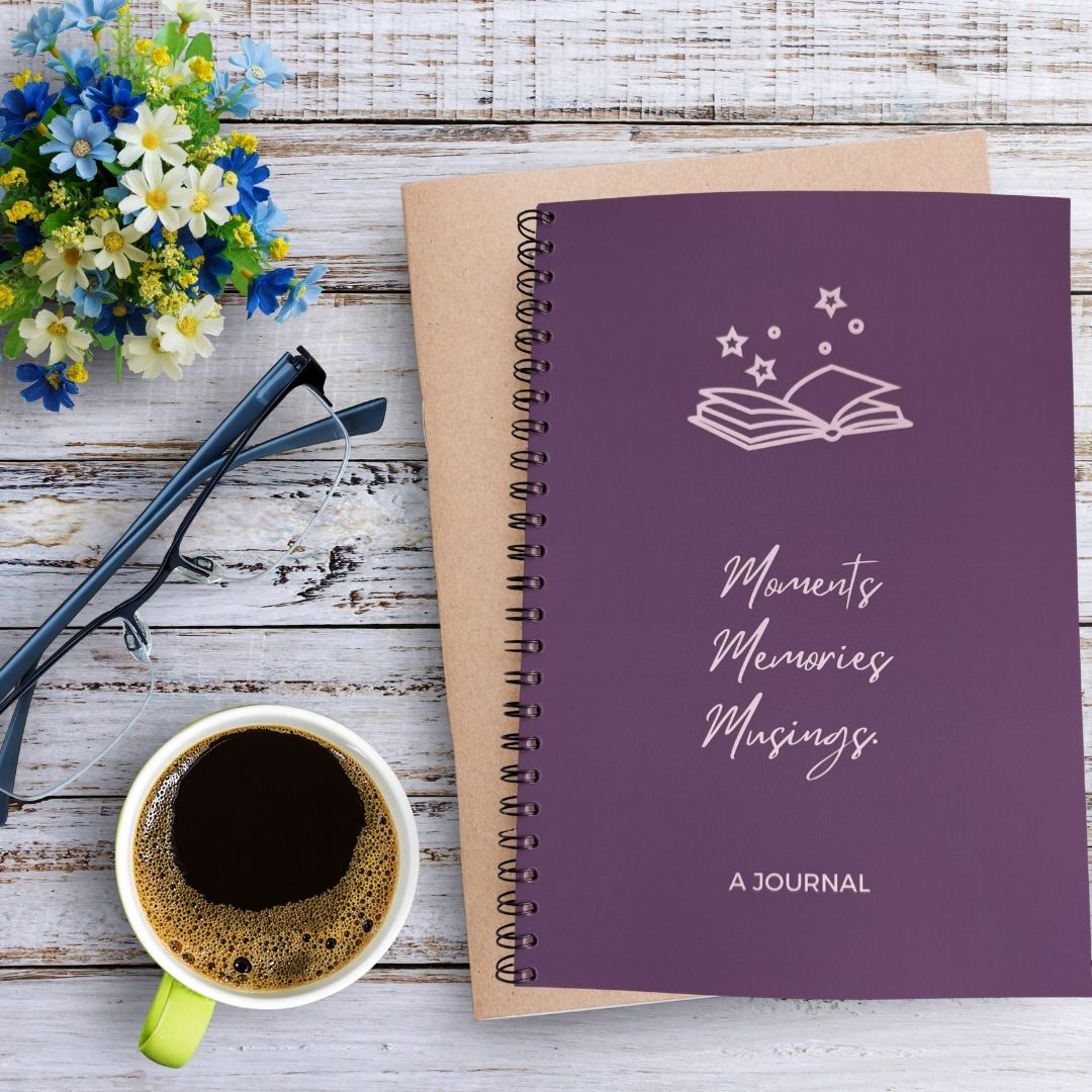 A journal with a solid plum purple background and white line art of an open book with stars above the title 'Moments Memories Musings' and 'A JOURNAL' text at the bottom. The journal sits on a bleached wooden table besides a mug of coffee, pair of glasses, and blue white green flower arrangement.