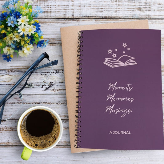 A journal with a solid plum purple background and white line art of an open book with stars above the title 'Moments Memories Musings' and 'A JOURNAL' text at the bottom. The journal sits on a bleached wooden table besides a mug of coffee, pair of glasses, and blue white green flower arrangement.