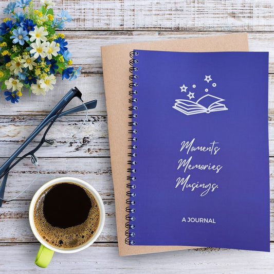 A journal with a solid deep blue background and white line art of an open book with stars above the title 'Moments Memories Musings' and 'A JOURNAL' text at the bottom. The journal sits on a bleached wooden table besides a mug of coffee, pair of glasses, and blue white green flower arrangement.