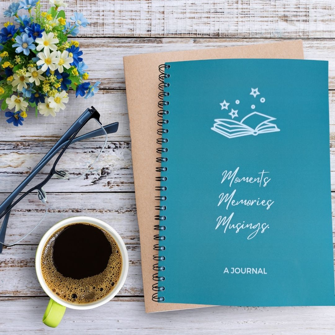 A teal spiral-bound journal with a white book-and-stars illustration and the words 'Moments Memories Musings' on the cover, styled on a whitewashed wood surface with a cup of coffee, eyeglasses, and small flowers.