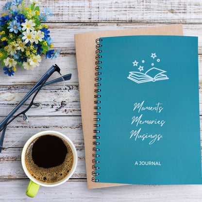 A teal spiral-bound journal with a white book-and-stars illustration and the words 'Moments Memories Musings' on the cover, styled on a whitewashed wood surface with a cup of coffee, eyeglasses, and small flowers.