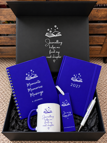 A curated gift set in an open black box with a dark message printed inside, featuring a wirebound blue journal, a blue diary, a white mug with blue accents, a blue bookmark, and a white pen. The journals and mug are printed with white book-and-stars graphics and the phrase 'Journalling helps me find my next chapter'.