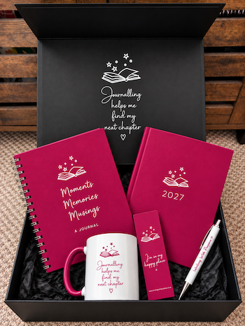 A curated journalling gift set displayed inside an open black box. The set includes a magenta spiral-bound journal, a magenta diary with '2027' printed on the cover, a white ceramic mug with magenta text and handle, a magenta bookmark, and a white pen with magenta lettering.