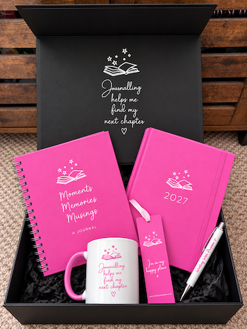 A curated self-care gift set in an open black box, featuring a pink spiral-bound journal, a pink 2027 diary, a white mug with pink text, a pink bookmark, and a white pen with pink detailing. The box lid is printed with a white illustration of an open book and the quote 'Journalling helps me find my next chapter'.