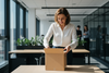 Create an image that shows a square cardboard box on a table in an office setting with a lady in her 40s, dressed in a blouse opening the top of the box, looking inside.